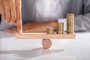  Close-up Of Businessperson's Finger Balancing Stacked Of Coins On Wooden SeesawIndexador: Andrey PopovFonte: 190662072<!-- NICAID(14394281) -->
