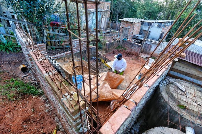 Sem dinheiro para contratar serventes, amarra os ferros das vigas, mistura a argamassa e coloca as pedras para a base da construção