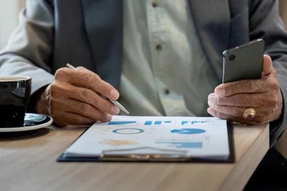 Senior Analysis and Communication. A senior man reviewing business charts while holding a smartphone in a cafe.An elderly man examines financial trends on a printed chart, taking notes with a pen, alongside a cup of coffee, highlighting focused analysis and planning.Fonte: 1334286328<!-- NICAID(16137755) -->
