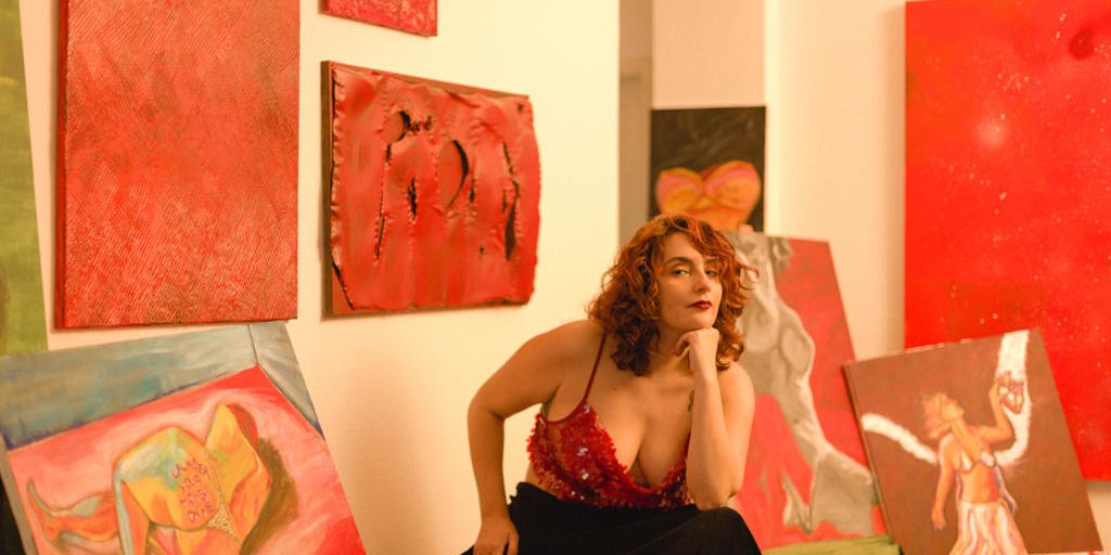 Bruna Maia é uma mulher branca que está sentada no centro da foto, cercada por telas que ela pintou. Tem nus, um mulher segurando o próprio coração, quadros inteiramente vermelhos.