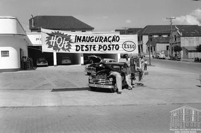 Inauguração do Posto Gazolube em 1953. Texto: Tendo como um dos principais sócios o empresário e piloto de corridas Oscar Bay, o Posto de Serviços Gazolube ganhou uma badalada inauguração em 5 de fevereiro de 1953. Localizado na esquina das ruas Os Dezoito do Forte e Marechal Floriano, o empreendimento da Companhia Esso também serviu de vitrine para o Jeep Willys Overland 1953, revendido pela Concessionária Bay & Cia Ltda.Conforme destacado na edição de 8 de fevereiro de 1953 do jornal A Época, o estabelecimento dispunha de duas bombas de gasolina elétricas, com tanques com capacidade para 35 mil litros, além de uma bomba de óleo diesel para 10 mil litros. Já os serviços de lavagem e lubrificação dividiram-se em três seções: a de caminhões, a de automóveis e a de lavagem da parte externa dos veículos.Na foto: momento da solenidade de inauguração, da qual participaram Oscar Bay, padre Gasparotto, Nadyr Festugato, Darvile Croda, Artemin Karam, fornecedores, diretores da Esso e funcionários do posto.<!-- NICAID(16046695) -->