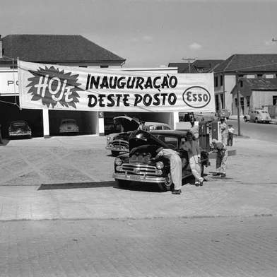 Inauguração do Posto Gazolube em 1953. Texto: Tendo como um dos principais sócios o empresário e piloto de corridas Oscar Bay, o Posto de Serviços Gazolube ganhou uma badalada inauguração em 5 de fevereiro de 1953. Localizado na esquina das ruas Os Dezoito do Forte e Marechal Floriano, o empreendimento da Companhia Esso também serviu de vitrine para o Jeep Willys Overland 1953, revendido pela Concessionária Bay & Cia Ltda.Conforme destacado na edição de 8 de fevereiro de 1953 do jornal A Época, o estabelecimento dispunha de duas bombas de gasolina elétricas, com tanques com capacidade para 35 mil litros, além de uma bomba de óleo diesel para 10 mil litros. Já os serviços de lavagem e lubrificação dividiram-se em três seções: a de caminhões, a de automóveis e a de lavagem da parte externa dos veículos.Na foto: momento da solenidade de inauguração, da qual participaram Oscar Bay, padre Gasparotto, Nadyr Festugato, Darvile Croda, Artemin Karam, fornecedores, diretores da Esso e funcionários do posto.<!-- NICAID(16046695) -->