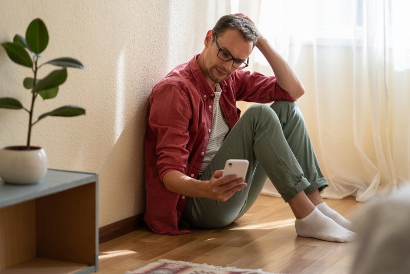 Upset young man holding smartphone waiting for ex-girlfriend call while sitting on floor at homeHomem esperando mensagem - Foto: DimaBerlin/stock.adobe.comFonte: 545688027<!-- NICAID(15743264) -->