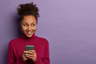 Smiling pleasant looking woman receives message from dating app, holds mobile phone in hands, looks happily aside, edits photo to post online, has curly hair, isolated on violet wall, copy spaceMulher com celular na mão - Foto: Wayhome Studio/stock.adobe.comFonte: 333676070<!-- NICAID(15377191) -->