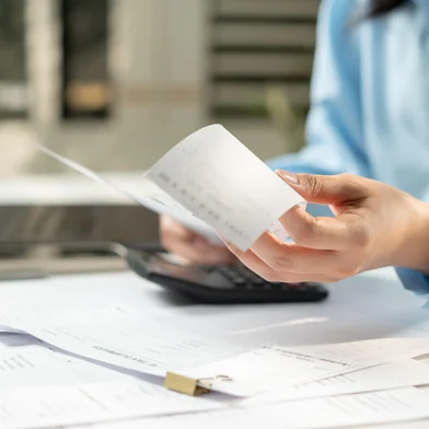 Businesswoman hands review receipts beside laptop and calculator. Focused expression shows careful financial management. Desk cluttered with papers implies busy work environment.<!-- NICAID(16339767) -->