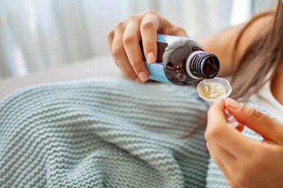 Anonymous sick female filling measuring spoon with cough syrup while sitting under blanket at home. Girl drinks a medicine. She is being treated for a cough. Treatment of influenza.Fonte: 368938548<!-- NICAID(16033974) -->