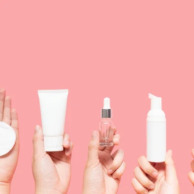 Skincare routine step for healthy skin - Woman hands holding facial cotton pad, foam, essential oil, serum, lotion and eye cream packaging on pink background. Beauty and cosmetic concept. Copy space.<!-- NICAID(16122648) -->