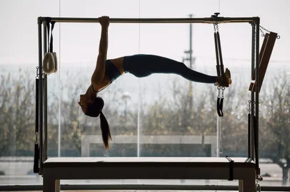Fit female coach hanging hands and feet from the Pilates machine, woman working out on the Trapeze Table in a big Fitness Gym, big windows and street views. Pilates and healthy lifestyle conceptFonte: 419365360<!-- NICAID(15673409) -->