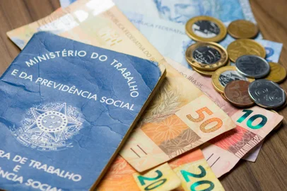 Notas de dinheiro acompanhado de moedas, carteira de trabalho, lupa, calculadora encima da mesa. Foto: fernando / stock.adobe.comIndexador: PEDROFonte: 186683303<!-- NICAID(15400603) -->