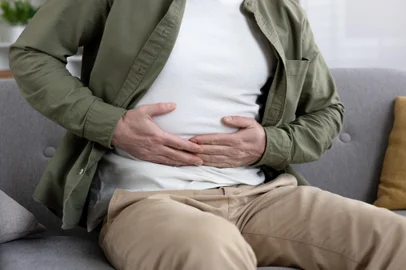 Man experiencing abdominal pain while sitting on couch holding stomach in discomfortMan sitting on couch experiencing abdominal pain holding stomach. Wearing casual clothing and showing visible discomfort. Concept of stomachache, digestive issues, or health concerns.Indexador: Liubomyr VoronaFonte: 931328034<!-- NICAID(15948898) -->