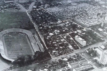 A imagem aérea acima integrou a exposição fotográfica Euzébio Beltrão de Queiróz Favela do Cemitério, realizada em 1984 na administração do prefeito Victório Trez. A mostra contou originalmente com 29 quadros, dotados de imagens e textos sobre o bairro: histórico, situação e localização, topografia, pavimentação e sistema viário, rede de água e esgoto, coleta de lixo, e equipamento público e social. Em relação aos textos, destacamos: No início da década de 50, Getúlio Vargas visita a área onde havia 50 habitações, sendo a única favela dessa dimensão no município. Em 1963, foi feito um cadastramento e foram identificadas 201 casas, com 986 habitantes, numa área de 3,5 ha. A divisa do cemitério era de cerca viva. A partir desta data foi construído o muro de alvenaria e a área diminuiu para 2,5 ha.Viabilizada pelos arquitetos Celito Girardi, João Batista Mosele, Juarez Marchioro, Maria Conceição Baldisserotto, Paula Folchini, sob a assessoria do arquiteto Antonio Fillipini, as imagens fotográficas da exposição são de autoria de Antônio Carlos Galvão e Maria Conceição Baldisserotto. A exposição completa está disponível em: arquivomunicipal.caxias.rs.gov.br.<!-- NICAID(15445891) -->