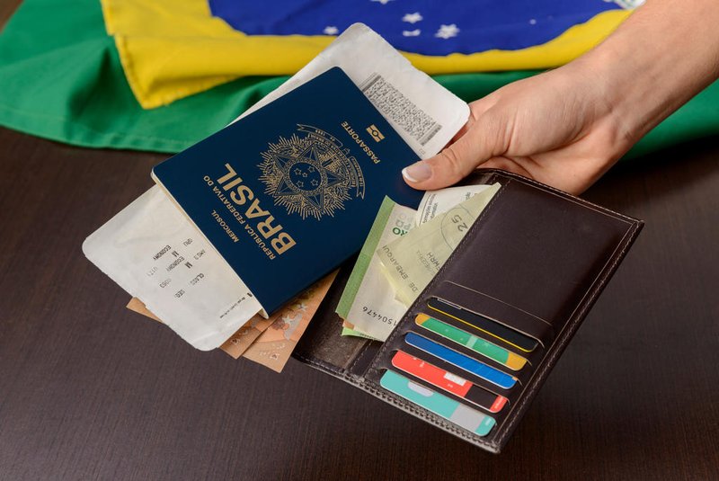Hand holding Brazilian passport over table with Brazilian flag in the background.*A PEDIDO DE GABRIELA PERUFO* Hand holding Brazilian passport over table with Brazilian flag in the background. - Foto: Cacio Murilo/stock.adobe.comIndexador: Cacio Murilo de VasconcelosFonte: 523946492Fotógrafo: FotÃ³grafo<!-- NICAID(15195610) -->