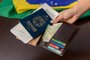 Hand holding Brazilian passport over table with Brazilian flag in the background.*A PEDIDO DE GABRIELA PERUFO* Hand holding Brazilian passport over table with Brazilian flag in the background. - Foto: Cacio Murilo/stock.adobe.comIndexador: Cacio Murilo de VasconcelosFonte: 523946492Fotógrafo: FotÃ³grafo<!-- NICAID(15195610) -->