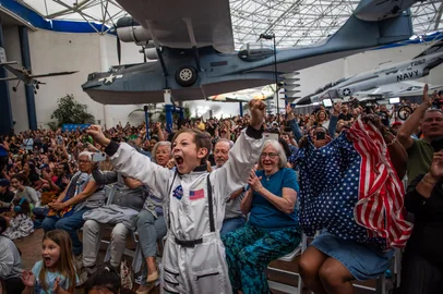 A young boy wearing an astronaut costume cheers next to a woman waving a flag as they watch a live broadcast of the return of the Artemis II crew members to Earth at the San Diego Air and Space Museum during a watch party for the crew's splash down in the Pacific Ocean, in San Diego, California, on April 10, 2026. The NASA spacecraft carrying four astronauts splashed down as planned Friday off the California coast, capping the US space agency's successful crewed test mission around the Moon, the first such flyby in more than 50 years. Mission commander Reid Wiseman reported that the crewmembers -- himself along with Christina Koch, Victor Glover and Jeremy Hansen -- were "stable" and "green." (Photo by Apu GOMES / AFP)<!-- NICAID(16263830) -->