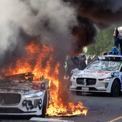 Demonstrators smash the windshield of a vehicle next to a burning Waymo vehicle as protesters clash with law enforcement in the streets surrounding the federal building during a protest following federal immigration operations in Los Angeles, California, on June 8, 2025. Demonstrators torched cars and scuffled with security forces in Los Angeles on June 8, as police kept protestors away from the National Guard troops President Donald Trump sent to the streets of the second biggest US city. (Photo by RINGO CHIU / AFP)<!-- NICAID(16055364) -->
