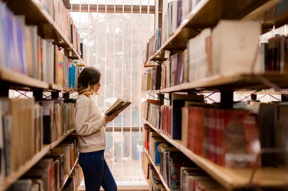 Cute  girl reading a book while standing between the bookshelves. Foto: qunica.com / stock.adobe.comFonte: 529150641<!-- NICAID(16090563) -->