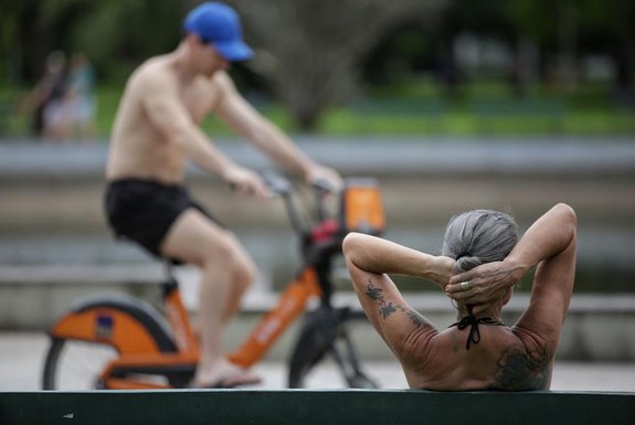 Mulher sentada em banco no Parque da Redenção, ao fundo, um homem passa em uma bicicleta laranja do Itaú.<!-- NICAID(15615146) -->