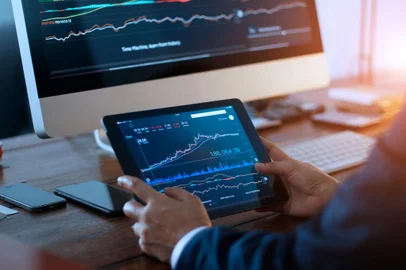 Businessman checking stock market on digital tablet and a desktop computer with stock exchange graph on screen. Financial stock market. Analyzing data in office background.Fonte: 268788365<!-- NICAID(14892507) -->