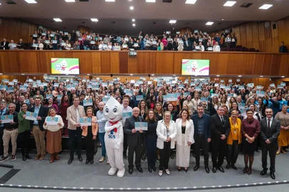 Auditório amplo e lotado, com duas galerias, repleto de pessoas em pé segurando certificados azuis voltados para a câmera. Na frente, um grupo de autoridades posa para a foto, ao lado do mascote Zé Gotinha, símbolo das campanhas de vacinação do SUS. As paredes de madeira e o teto com iluminação clara compõem o ambiente. Ao fundo, dois telões exibem a imagem do Zé Gotinha sobre fundo colorido, reforçando a temática de saúde e vacinação.<!-- NICAID(16106099) -->