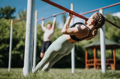 Two individuals perform calisthenics exercises in an urban park, utilizing outdoor bars on a sunny day, promoting fitness and a healthy lifestyle.<!-- NICAID(16237938) -->