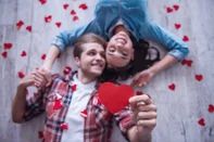 Top view of happy young couple looking at camera and smiling while lying on wooden floor. Guy is holding a red paper heart<!-- NICAID(16304697) -->