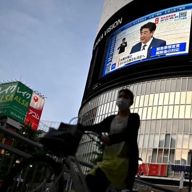 Japan's Prime Minister Shinzo Abe (top R) is seen on a live TV broadcast as he announces the lifting of a nationwide state of emergency, as a woman on a bicyle crosses the road in Tokyo’s Shinjuku area on May 25, 2020. - Japan Prime Minister Shinzo Abe on May 25 lifted the state of emergency imposed nationally to combat coronavirus following a sharp decline in the number of new cases. (Photo by CHARLY TRIBALLEAU / AFP)