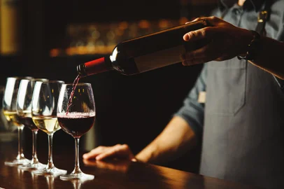 Bartender pours red wine in glasses on wooden bar counterBartender in apron pours sweet red wine from bottle in capacious glasses on wooden bar counter. Delicious alcohol drink in transparent glassware with nice scent.Fonte: 188192171<!-- NICAID(15306148) -->