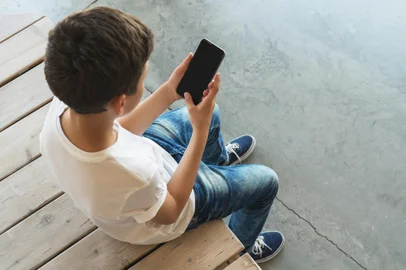View from above. Boy in white T-shirt and sunglasses sitting indoor and uses smartphone. Teenager plays computer gamesConceito de jovem, adolescente, criança no celular - Foto: foxyburrow/stock.adobe.comFonte: 183672881<!-- NICAID(15902314) -->