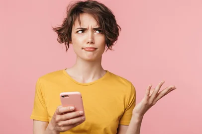 Confused young beautiful woman posing isolated over pink wall background using mobile phone.Imagem de uma mulher seurando o telefone. Foto: Drobot Dean / stock.adobe.comFonte: 271731872<!-- NICAID(15065383) -->