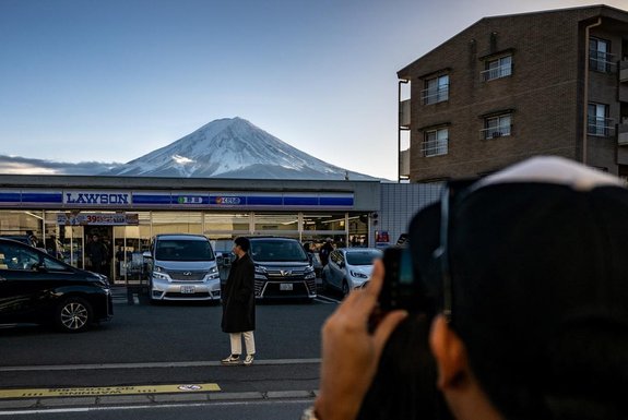 This photo taken on January 1, 2024 shows a tourist posing in front of a convenience store with Mount Fuji in the background, in the town of Fujikawaguchiko, Yamanashi prefecture. A huge black barrier to block Mount Fuji from view will be installed in a popular photo spot by Japanese authorities exasperated by crowds of badly behaved foreign tourists, it was reported on April 26, 2024. (Photo by Philip FONG / AFP)Editoria: LIFLocal: FujikawaguchikoIndexador: PHILIP FONGSecao: mountainsFonte: AFPFotógrafo: STF<!-- NICAID(15745823) -->
