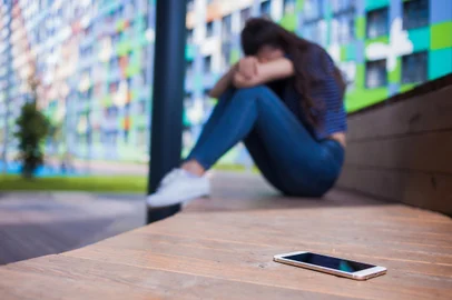Smartphone lying in the foreground, on blurred background, girl, hiding face in her knees, sitting on a wooden bench.A smartphone lying in the foreground, on a blurred background, a girl, a brunette with long hair in jeans and white sneakers, crying, hiding her face in her knees, sitting with her legs on a wooden bench. Side view.Fonte: 280582321<!-- NICAID(15848028) -->