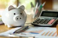 A piggy bank sits next to a calculator and financial charts on a wooden desk, symbolizing personal savings and financial planning.Conceito de dinheiro, economia, finanças, poupança - Foto: MP Studio/stock.adobe.comIndexador: mikeaistockFonte: 749184941<!-- NICAID(15893745) -->