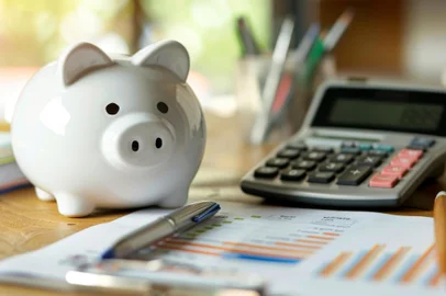 A piggy bank sits next to a calculator and financial charts on a wooden desk, symbolizing personal savings and financial planning.Conceito de dinheiro, economia, finanças, poupança - Foto: MP Studio/stock.adobe.comIndexador: mikeaistockFonte: 749184941<!-- NICAID(15893745) -->