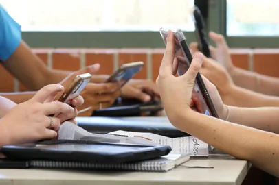 A table of high school students all holding multiple digital devices such as phones and tablets during a class lesson. Contemporary education and connected technology. No identifiable people or property visible. - Foto: LincB/stock.adobe.comFonte: 390249107<!-- NICAID(15348165) -->