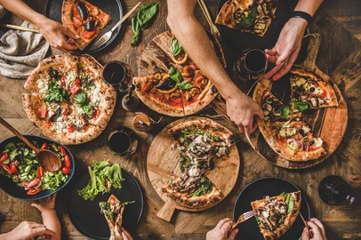People eating different kinds of pizza, salad and drinking wineFamily or friends having pizza party dinner. Flat-lay of people eating different kinds of Italian pizza, salad and drinking wine over wooden table, top view. Fast food lunch, gathering, celebration. Foto: sonyakamoz / stock.adobe.comFonte: 308455555<!-- NICAID(16076424) -->