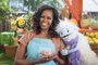Michelle Obama, wearing a blue apron, stands in a rooftop garden posing for the camera and holding a pink, round mochi puppet. A bee puppet wearing a red tie hovers over her shoulder while a furry white and blue puppet with frozen waffle ears embraces her side. <!-- NICAID(14725246) -->