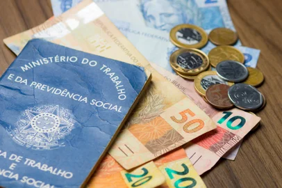 Notas de dinheiro acompanhado de moedas, carteira de trabalho, lupa, calculadora encima da mesa. Foto: fernando / stock.adobe.comIndexador: PEDROFonte: 186683303<!-- NICAID(15400603) -->