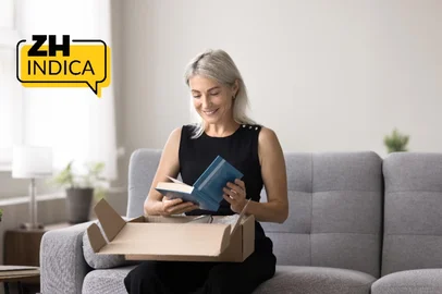 Smiling mature woman sitting on sofa unpacking box, get package, opening received parcel with ordered books using easy and comfortable on-line bookshop delivery services. Hobby, leisure, book lover<!-- NICAID(16198345) -->