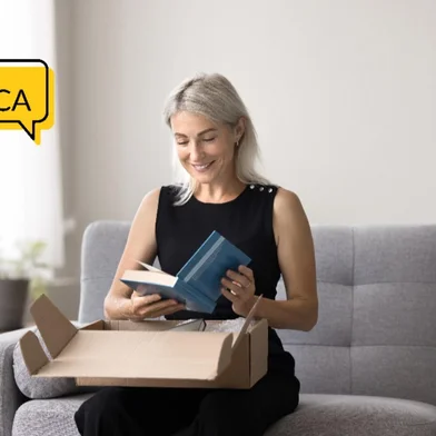 Smiling mature woman sitting on sofa unpacking box, get package, opening received parcel with ordered books using easy and comfortable on-line bookshop delivery services. Hobby, leisure, book lover<!-- NICAID(16198345) -->