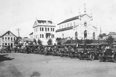 Pioneiros carros de praça de tração a motor estacionados na Rua Marquês do Herval, defronte à Praça Dante Alighieri, por volta de 1930. Entre as construções ao fundo, o antigo casarão e a casa de comércio da família Pezzi (atual Edifício Dona Ercília), o Bispado e a Catedral Diocesana, ainda com as escadarias frontais. Tudo em uma Caxias prestes a receber pavimentação.