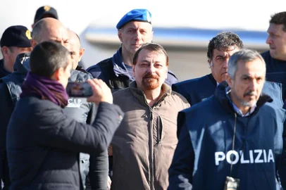  Italian former communist militant Cesare Battisti (C), wanted in Rome for four murders attributed to a far-left group in the 1970s, is escorted by Italian Police officers after stepping off a plane coming from Bolivia and chartered by Italian authorities, after landing at Ciampino airport in Rome on January 14, 2019. - Former communist militant Cesare Battisti landed in Rome on January 14 after an international police squad tracked the Italian down and arrested him in Bolivia, ending almost four decades on the run. (Photo by Alberto PIZZOLI / AFP)Editoria: POLLocal: RomeIndexador: ALBERTO PIZZOLISecao: politics (general)Fonte: AFPFotógrafo: STF
