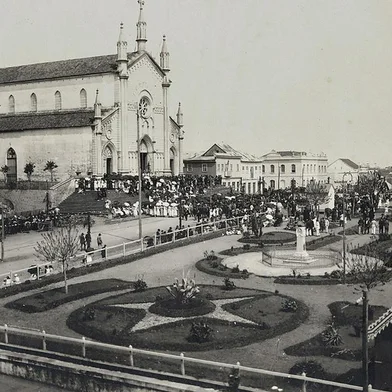 Quase não há fotos do centro de Caxias durante o surto de gripe espanhola, entre finais de 1918 e 1919, tampouco registros das medidas sanitárias junto aos vagões da Estação Férrea. O registro acima, do início da década de 1920, traz o cenário da Praça Dante Alighieri pós-epidemia, uma concentração de pessoas impensável dois anos antes, quando a cidade recolheu-se em casa. Trata-se de uma cerimônia religiosa captada a partir da Rua Marquês do Herval, com o recém-construído prédio do Bispado, a Catedral Diocesana, o casarão que abrigou a loja A Economia do Povo (embrião da Casa Magnabosco, de Raymundo Magnabosco) e o casarão de alvenaria da família Serafini (onde viveu a jovem Flora Serafini, filha do fotógrafo Giovanni Battista Serafini e esposa de Raymundo). Na praça ainda cercada por muros, o busto de Dante Alighieri, surgido em 1914 e envolto por grades (à frente), e o de Júlio Prates de Castilhos (ao fundo).<!-- NICAID(14468745) -->