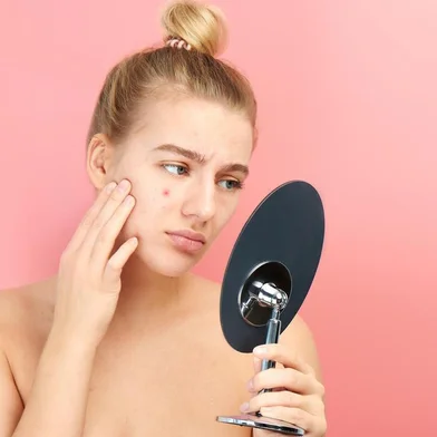 Upset girl looks in the mirror at her problem skin with red acne and post-acne isolated on a pink backgroundUpset girl looks in the mirror at her problem skin with red acne and post-acne isolated on a pink backgroundFonte: 302257784<!-- NICAID(14502572) -->