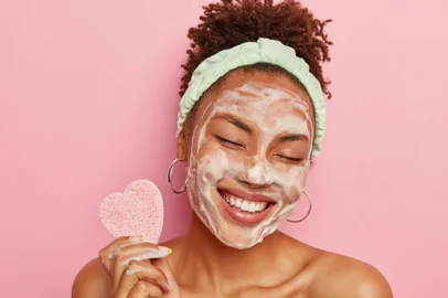 Beautiful optimistic Afro American woman cleanses face with foam, refreshes skin, has well cared complexion, holds heart shaped sponge for beauty procedures, stands bare shoulders with closed eyesBeautiful optimistic Afro American woman cleanses face with foam, refreshes skin, has well cared complexion, holds heart shaped sponge for beauty procedures, stands bare shoulders with closed eyesFonte: 288324777<!-- NICAID(14327345) -->