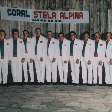 Uma das primeiras formações do grupo, em meados dos anos 1980, ainda com a denominação Coral Stela Alpina. Da esquerda para a direita estão Bortolo Menegol, Angelo Perini, Divino Pissaia, Arnaldo Pastori, Alziro Perini, Luiz Bertoni, Raimundo Sirena, José Demoliner, Armando Bertoni, José Comiotto, João Andreis, Honorino Rossa, Angelo Menegol, Domingos Dal Picol, Ledovino Isotton e Alcides Pissaia. <!-- NICAID(14581408) -->
