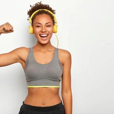 Horizontal view of dark skinned woman in good mood, raises arm with muscles, has strong body, dressed in gym outfit, listens audio via modern headphones, poses indoor. Fitness and music conceptVista horizontal da mulher de pele escura de bom humor, levanta o braço com músculos, tem corpo forte, vestido com roupa de ginástica, escuta áudio através de fones de ouvido modernos, poses indoor. Fitness e conceito de músicaFonte: 294064530<!-- NICAID(14316643) -->
