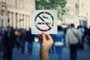 Hand holding a paper sheet with no smoking sign over a crowded street public place background. Forbidden area zone, restrictive symbol stop smoke.Fonte: 231724958<!-- NICAID(14866096) -->